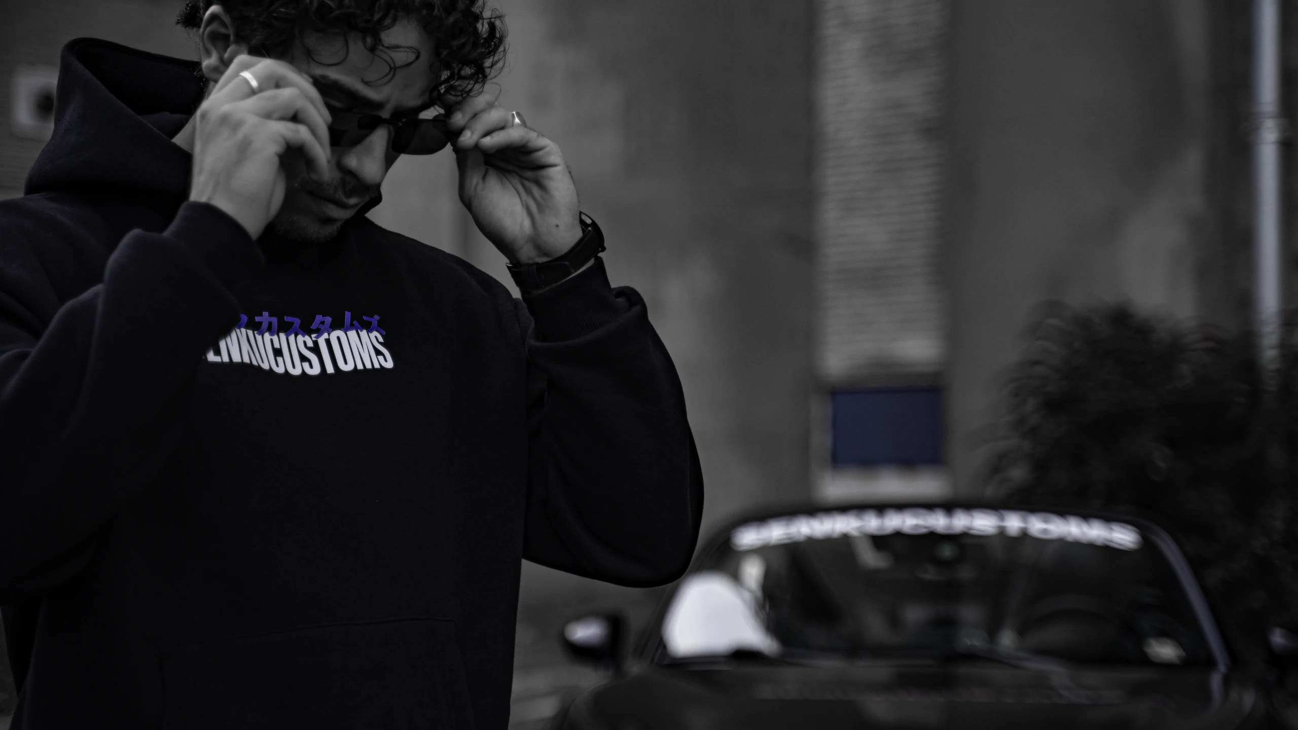 Man adjusting sunglasses in a black Senku Customs katakana hoodie, blurred MX-5 windscreen banner behind
