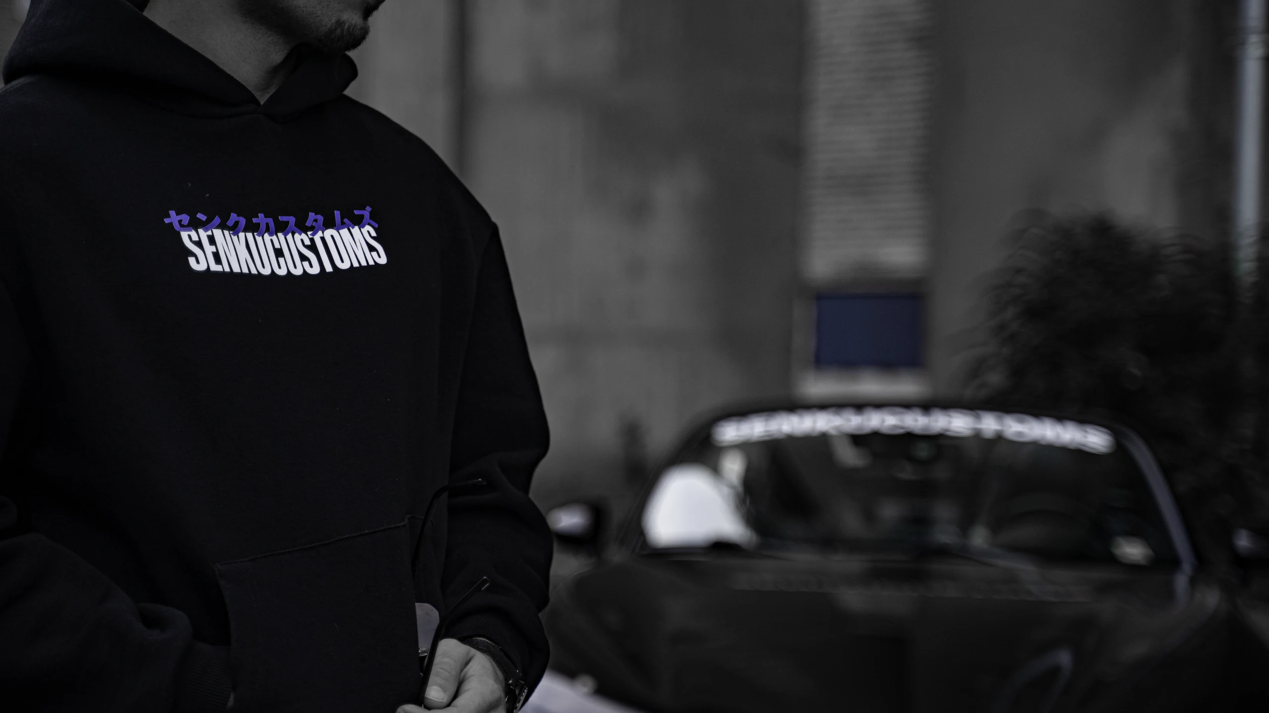Close-up of a black Senku Customs hoodie with purple katakana and white logo, blurred MX-5 behind