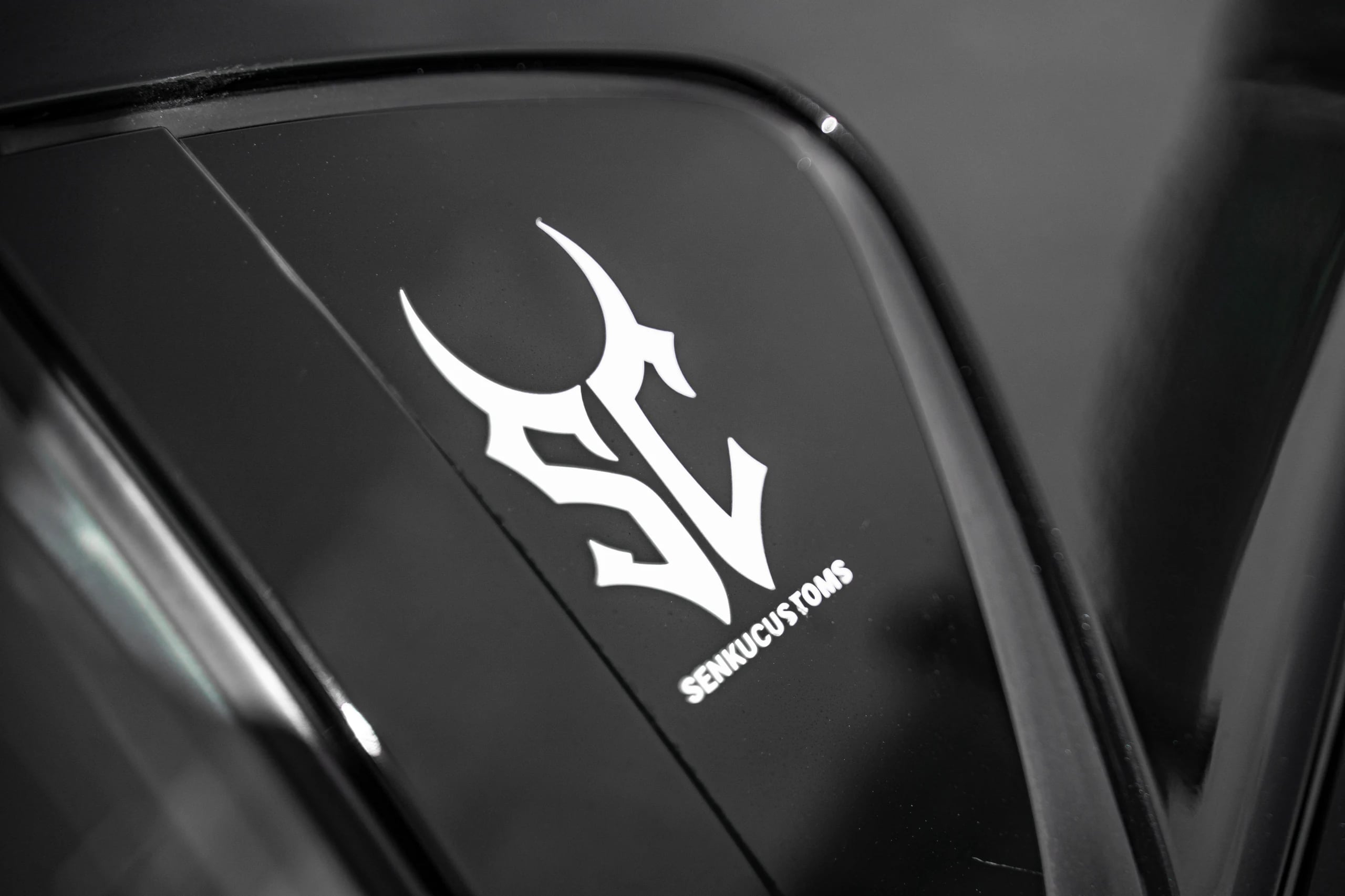 White Senku Customs SC logo decal on the gloss-black rear quarter window of a car, close-up